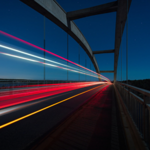 Shows a long exposure image of a car's rear lights going over a bridge